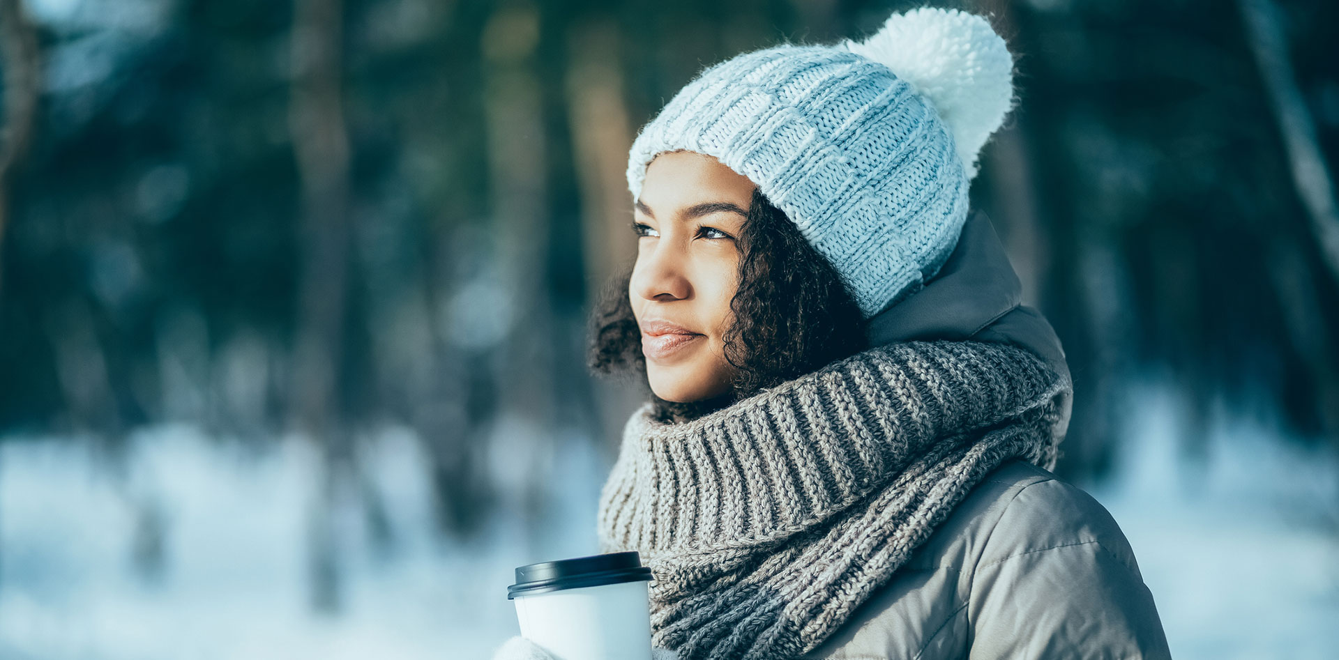 A young woman in a hat and mittens, holding a cup of coffee, outside in winter